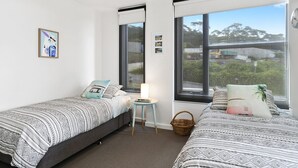 A bedroom with two single beds, a small table, a lamp, and a view of the outdoors.