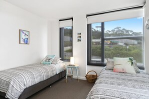 A bedroom with two single beds, a small table, a lamp, and a view of the outdoors.