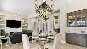 A dining area with a glass table, two chairs, a chandelier, and a kitchen with cabinets and a TV. - 3 Bedroom, PGA West home, Private Pool & Spa, #063306 (La Quinta)
