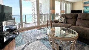 Een woonkamer met een bank, een glazen salontafel en een televisie. Er is een balkon met uitzicht op de oceaan en gebouwen.
