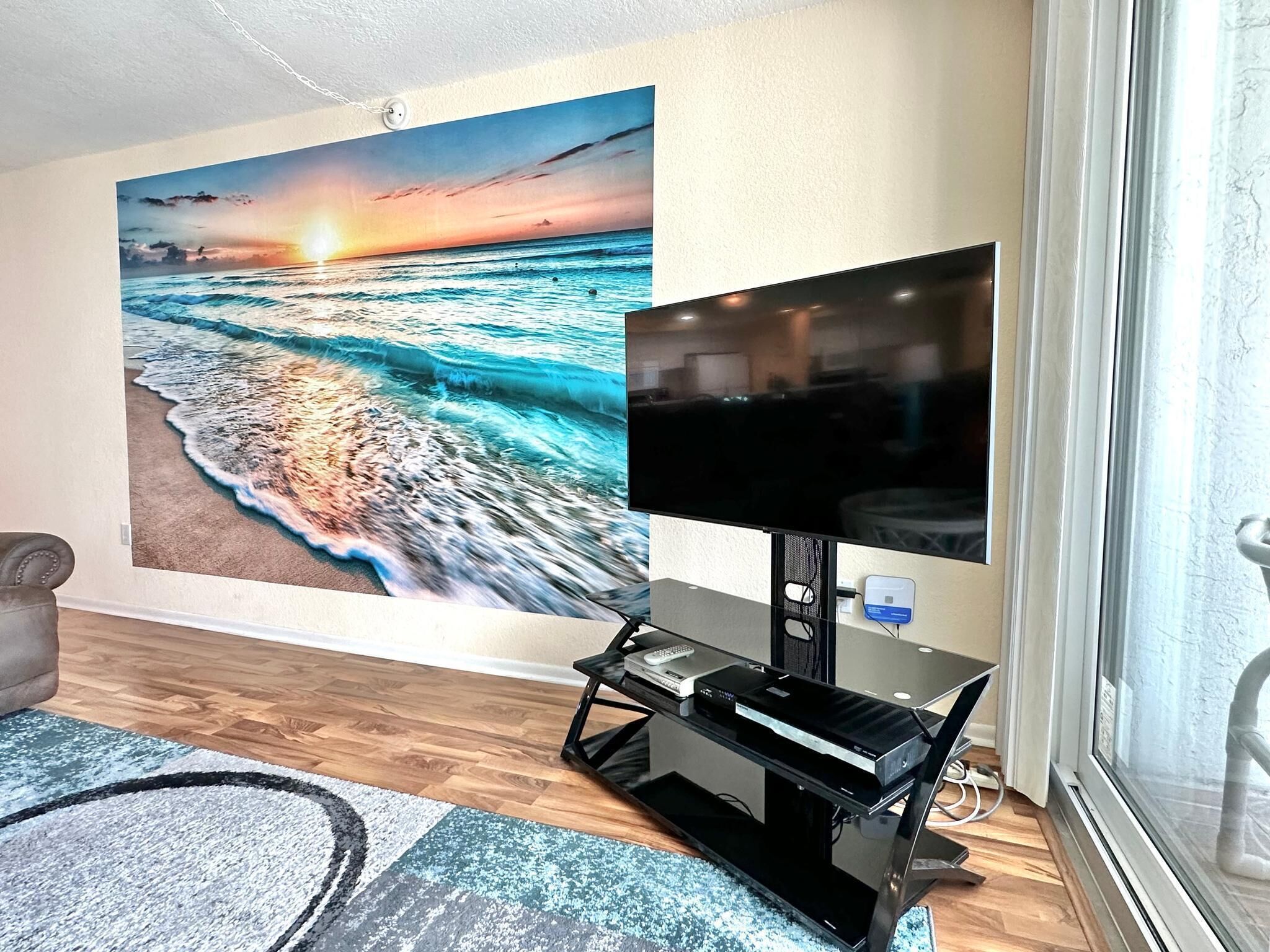 Una sala de estar con un gran mural de un atardecer sobre el océano, un televisor de pantalla plana sobre un soporte negro y un tapete azul estampado sobre el piso de madera.