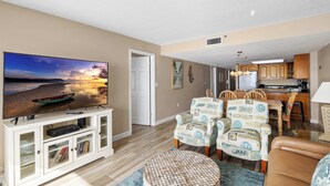 A living room with a TV, a dining area with chairs, and a kitchen in the background. - Direct Beachfront 3B2B Gulf - Beach Place #408 (Madeira Beach)