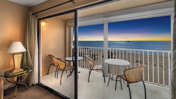 A balcony with a view of the ocean at sunset, furnished with wicker chairs and a table, and a lamp on the left. - Direct Beachfront 3B2B Gulf - Beach Place #408 (Madeira Beach)
