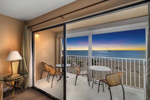 A balcony with a view of the ocean at sunset, furnished with wicker chairs and a table, and a lamp on the left.