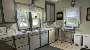 Une cuisine de taille réduite, avec des meubles blancs, un évier, une machine à café et une fenêtre ornée de rideaux en dentelle.