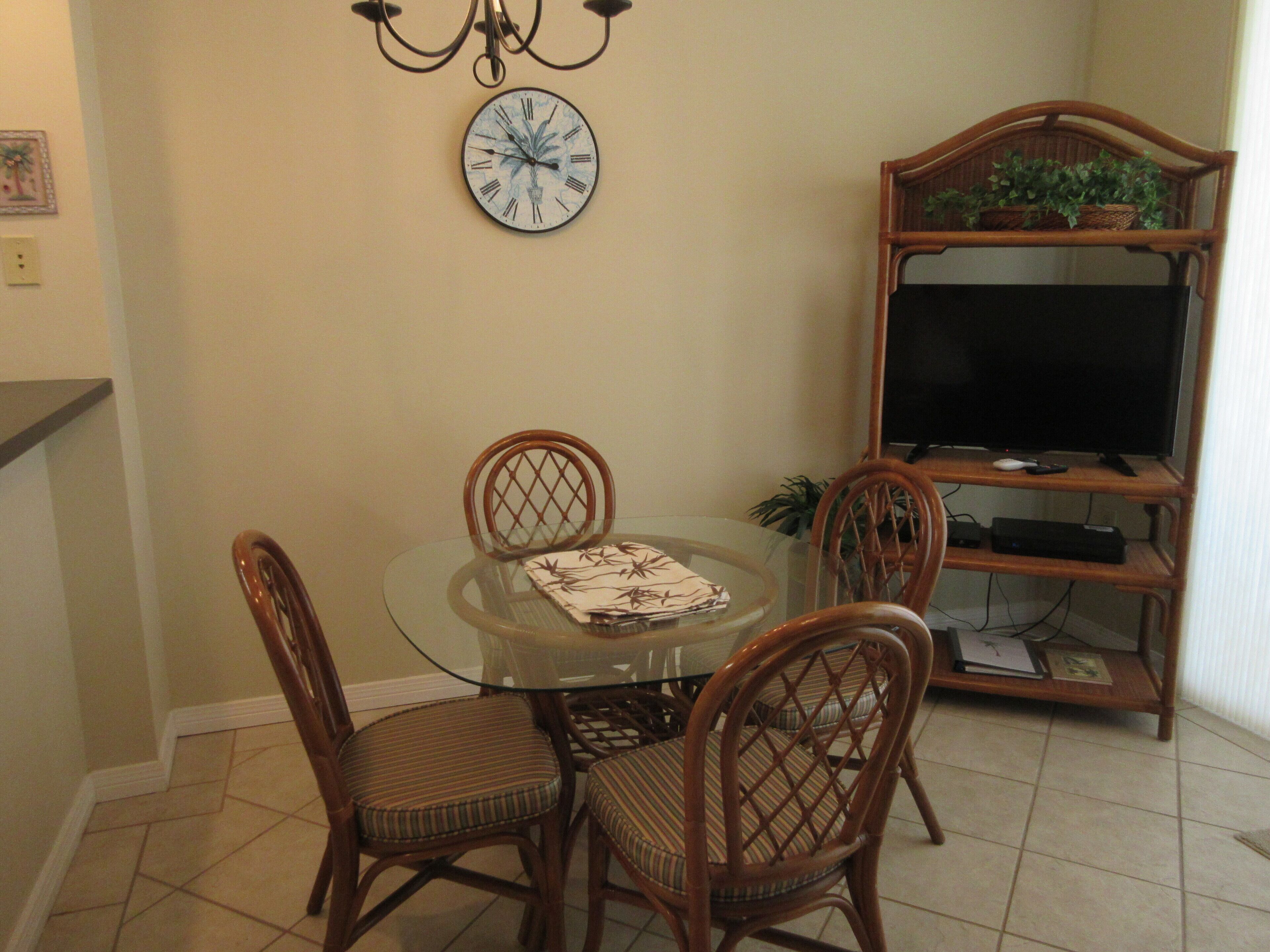 Un comedor con una mesa de vidrio, cuatro sillas de mimbre, un reloj en la pared y un televisor sobre un mueble de madera.