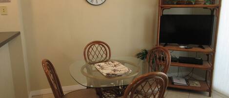 Un comedor con una mesa de vidrio, cuatro sillas de mimbre, un reloj en la pared y un televisor sobre un mueble de madera.