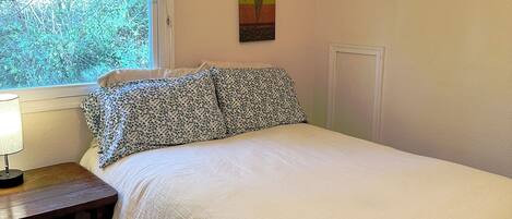 Quarto com cama, mesa de cabeceira, ventilador de teto, janela com persianas e um quadro na parede.