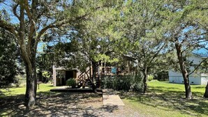 A pathway leading to a rustic wooden building with a porch, surrounded by trees and a gravel path. - McNeal Cottage:  Comfortable home, large private back yard. (Ocracoke)