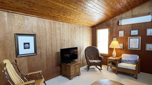A cozy room with wooden walls and ceiling, a television on a wooden stand, a rocking chair, a cushioned chair, a small table with a decorative item, and framed pictures on the wall. - McNeal Cottage:  Comfortable home, large private back yard. (Ocracoke)