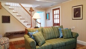A living room with a green sofa, wooden flooring, a staircase, and a door with a window. - The Mary Francis: Dog friendly, near Springers Point and Lighthouse (Ocracoke)