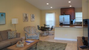 A living room with a sofa, a coffee table, a patterned armchair, a dining area with a table and chairs, and a kitchen with a refrigerator and cabinets.