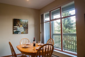 A dining area with a table set for four, a painting on the wall, and a large window with a view of trees. - Stoney Creek Northstar 36 (Whistler)