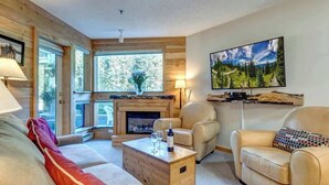 A cozy living room with a fireplace, leather sofas, a coffee table with wine glasses, and a large TV on the wall.