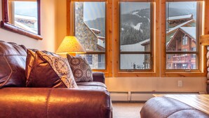 Un sofá de cuero con cojines estampados, una lámpara de mesa y grandes ventanas con vistas a un albergue de montaña.