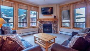 Una sala de estar acogedora con chimenea de piedra, un televisor de pantalla plana y grandes ventanales con vistas a una estación de esquí.