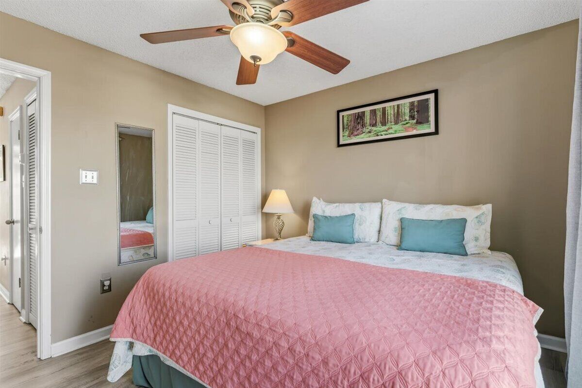 A bedroom with a bed, a ceiling fan, a door, a wall-mounted picture, and a nightstand with a lamp.