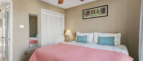 A bedroom with a bed, a ceiling fan, a door, a wall-mounted picture, and a nightstand with a lamp.