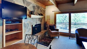 A living room with a stone fireplace, a flat-screen TV, and a balcony with a view of trees. - 🌲Timberline A2D Deluxe 2-Bedroom ~Hot Tub~Pool~WiFi~Ski-in/out~Local Shuttle🌲 (Snowmass Village)