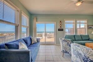 Une pièce avec un canapé bleu, un fauteuil à motifs et une vue sur la plage, visible à travers une porte-fenêtre coulissante.