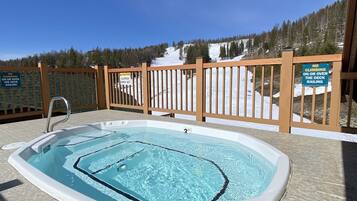 A hot tub with a wooden deck and railing, surrounded by a snowy landscape and trees.