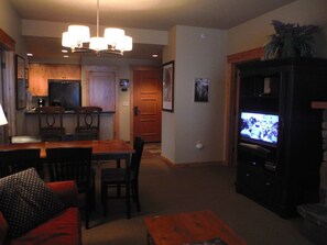 Ein Wohnzimmer mit Fernseher, ein Essbereich mit Stühlen und Tisch sowie eine Küche mit Schränken und Kühlschrank.