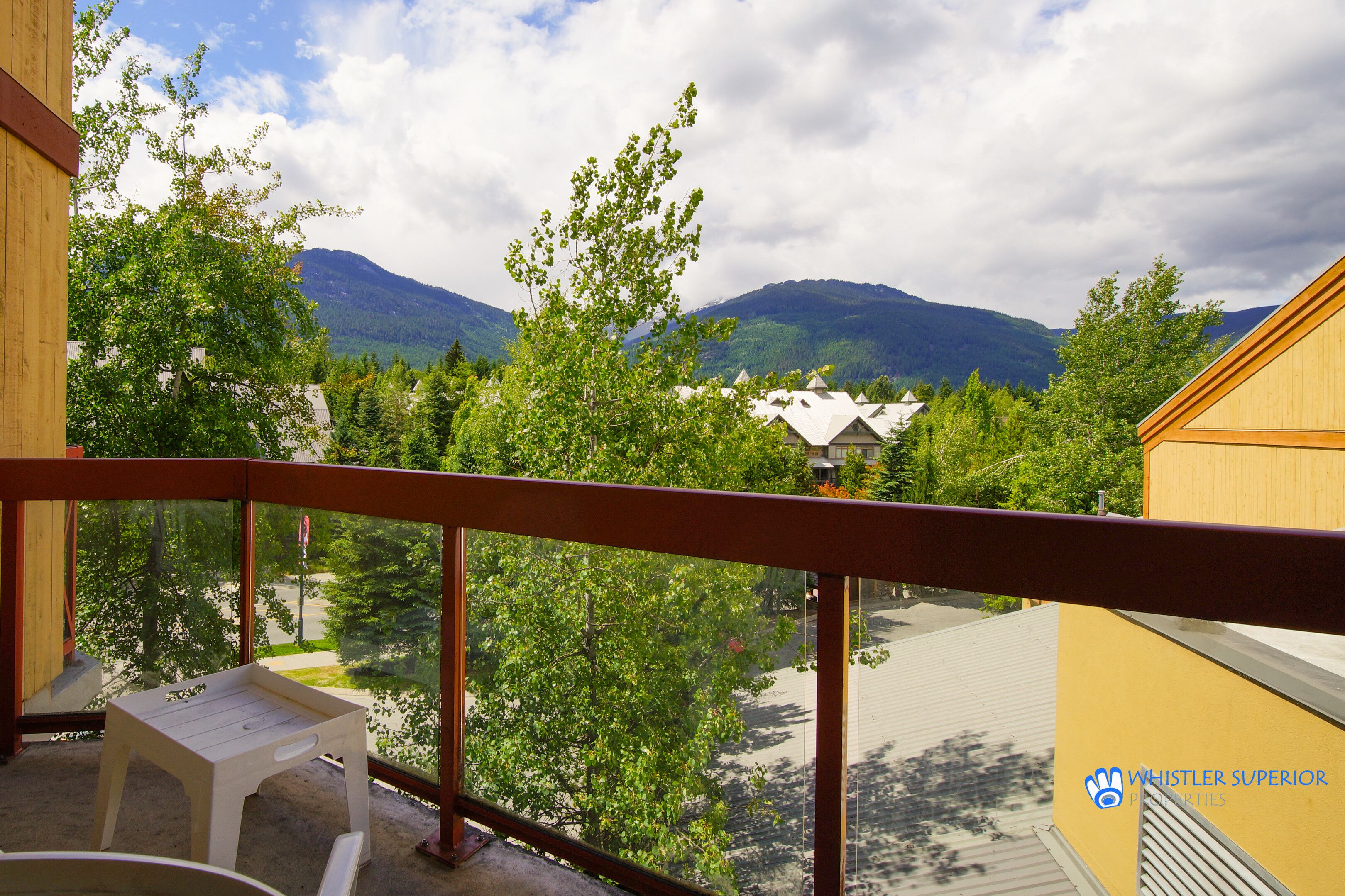 A balcony with a view of a mountain range, trees, and a building with a yellow facade.