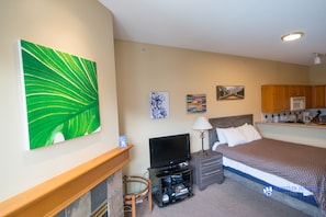 Une chambre d’hôtel avec un lit, une télévision, un bureau et une chaise. Il y a des tableaux au mur et, au fond, on aperçoit un coin cuisine.