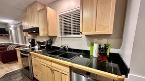 A modern kitchen with wooden cabinets, a black countertop, stainless steel appliances, and a window with blinds. - Cozy 2 Bedroom Waterfront Cottage with Hot Tub! (Ucluelet)