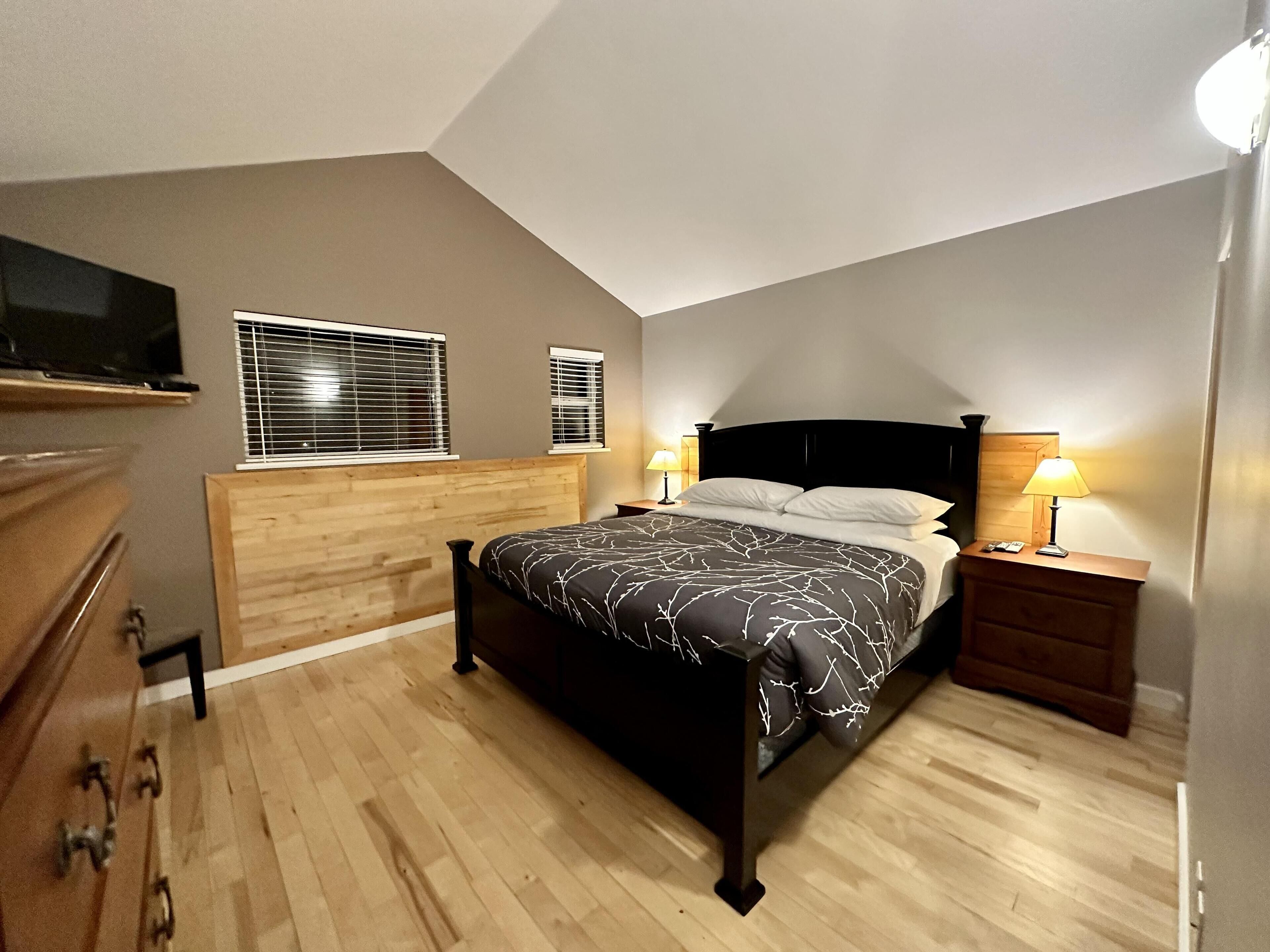 A bedroom with a wooden headboard, a bed with a patterned bedspread, two bedside lamps, and a television on a shelf above the bed.