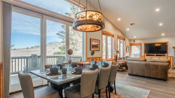 A dining area with a table set for six, large windows offering a view of the desert, a cozy living room with a sofa and TV, and a hanging light fixture. - Fantastic Mountain Townhome! Shared Hot Tub! Pool Table! Views! (Mammoth Lakes)