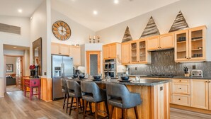 A modern kitchen with wooden cabinets, a stainless steel refrigerator, a central island with bar stools, and a decorative clock on the wall. - Fantastic Mountain Townhome! Shared Hot Tub! Pool Table! Views! (Mammoth Lakes)