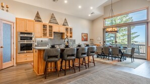 A spacious kitchen with wooden cabinets, a central island with bar stools, and a large dining area with a view of the outdoors. - Fantastic Mountain Townhome! Shared Hot Tub! Pool Table! Views! (Mammoth Lakes)