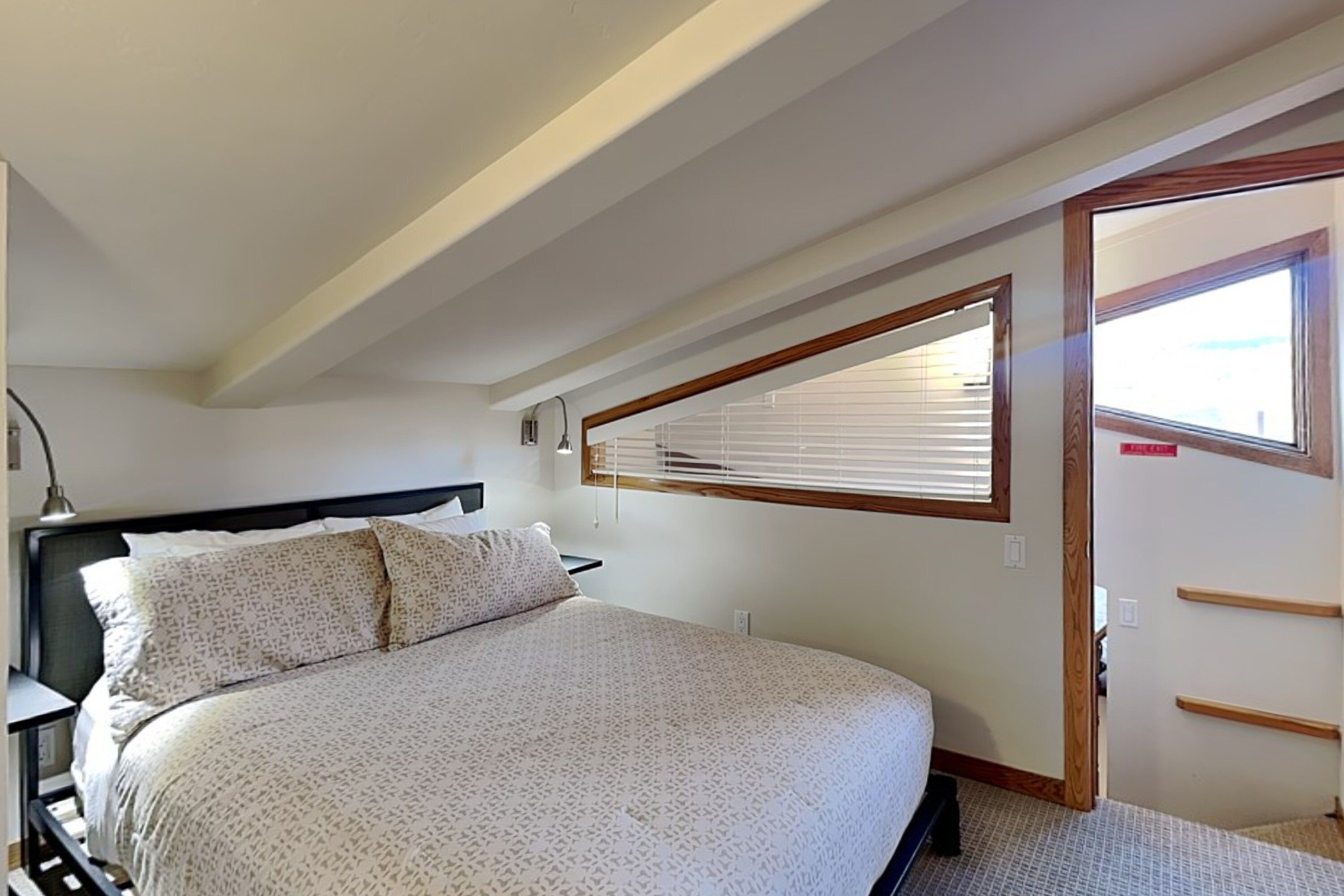 A bedroom with a bed, bedside tables, a window with blinds, and a door with a handle.