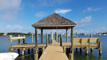 A wooden pier leading to a gazebo with a thatched roof, surrounded by calm water and a clear sky.