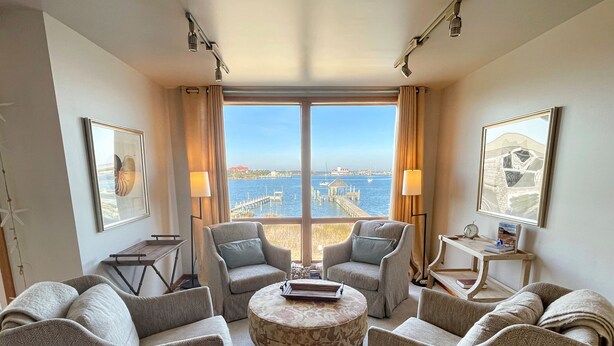 Ein Hotelzimmer mit großem Fenster und Blick auf das Meer und einen Steg. Es gibt zwei Sessel, einen runden Couchtisch und einen kleinen Beistelltisch mit Lampe.