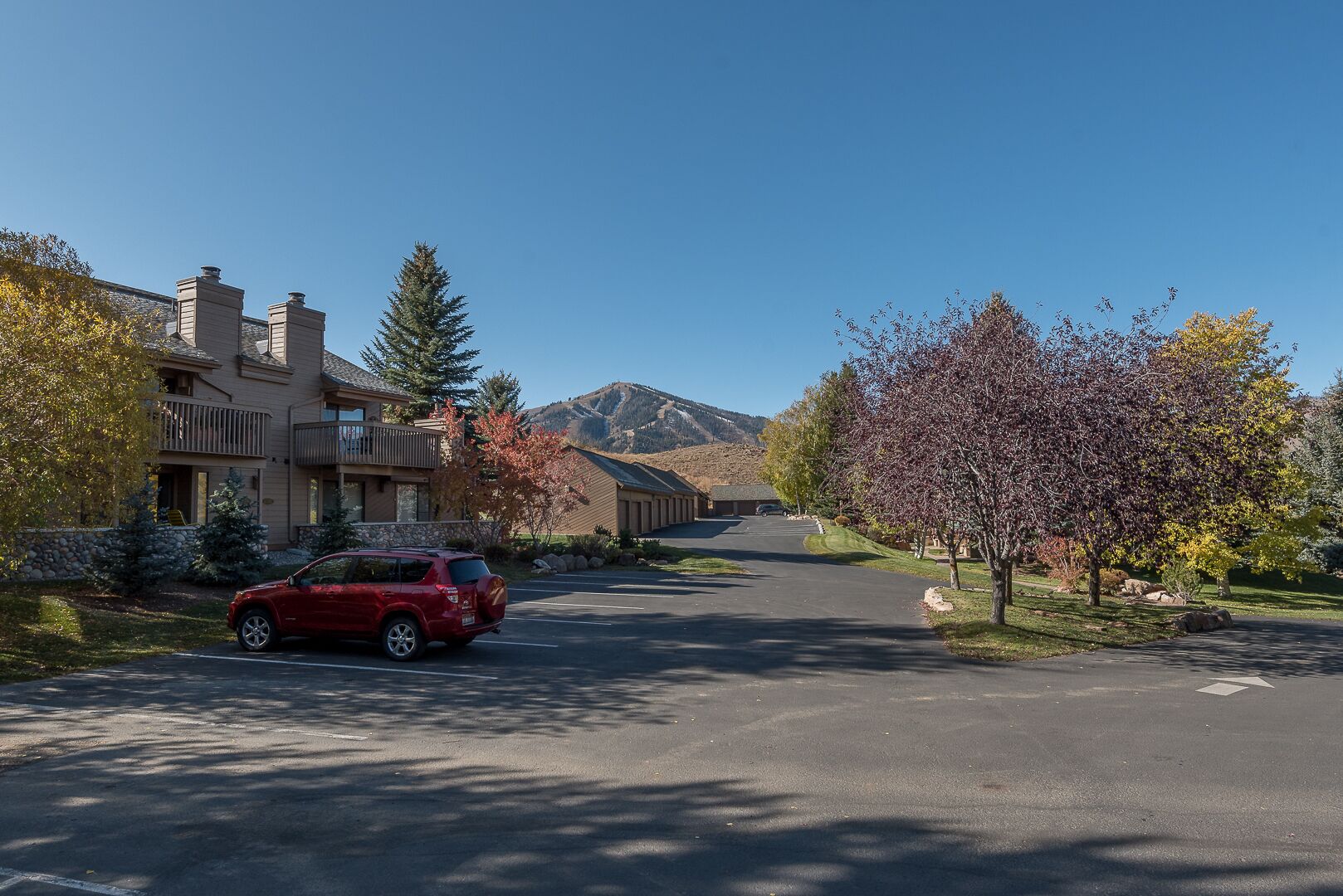 Une rue résidentielle avec un SUV rouge garé sur le côté gauche. On aperçoit des maisons avec des balcons et une montagne en arrière-plan.