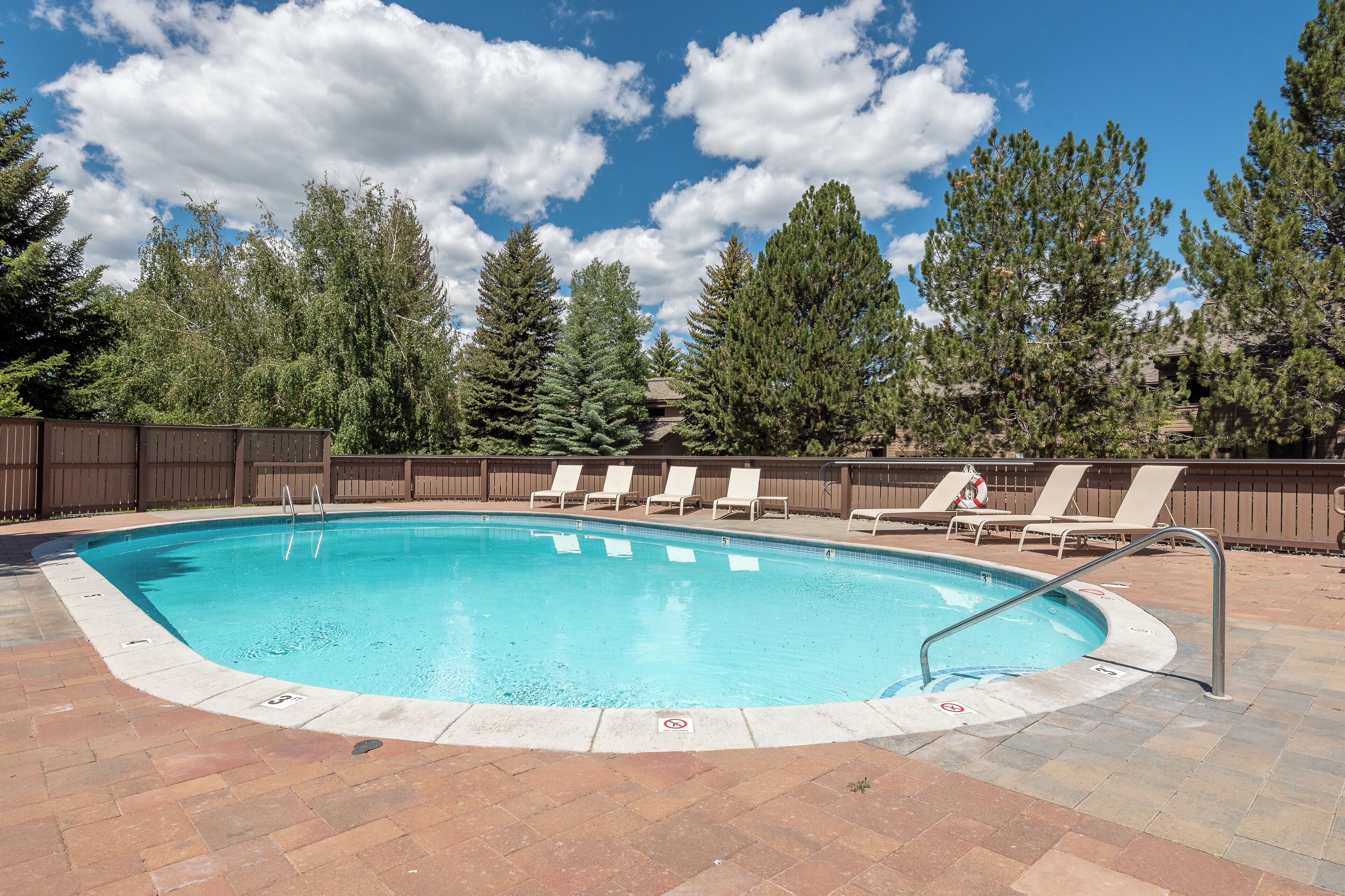 A rectangular swimming pool with a tiled deck, surrounded by lounge chairs and a wooden fence.