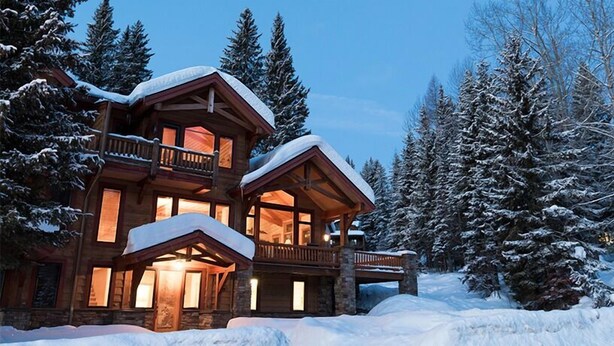Una cabaña de troncos con varios niveles, ventanas grandes y un balcón, rodeada de árboles cubiertos de nieve.