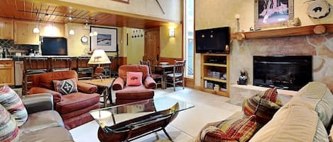 A spacious living room with leather sofas, a glass coffee table, and a fireplace. There is a kitchen area with wooden cabinets and a large TV.