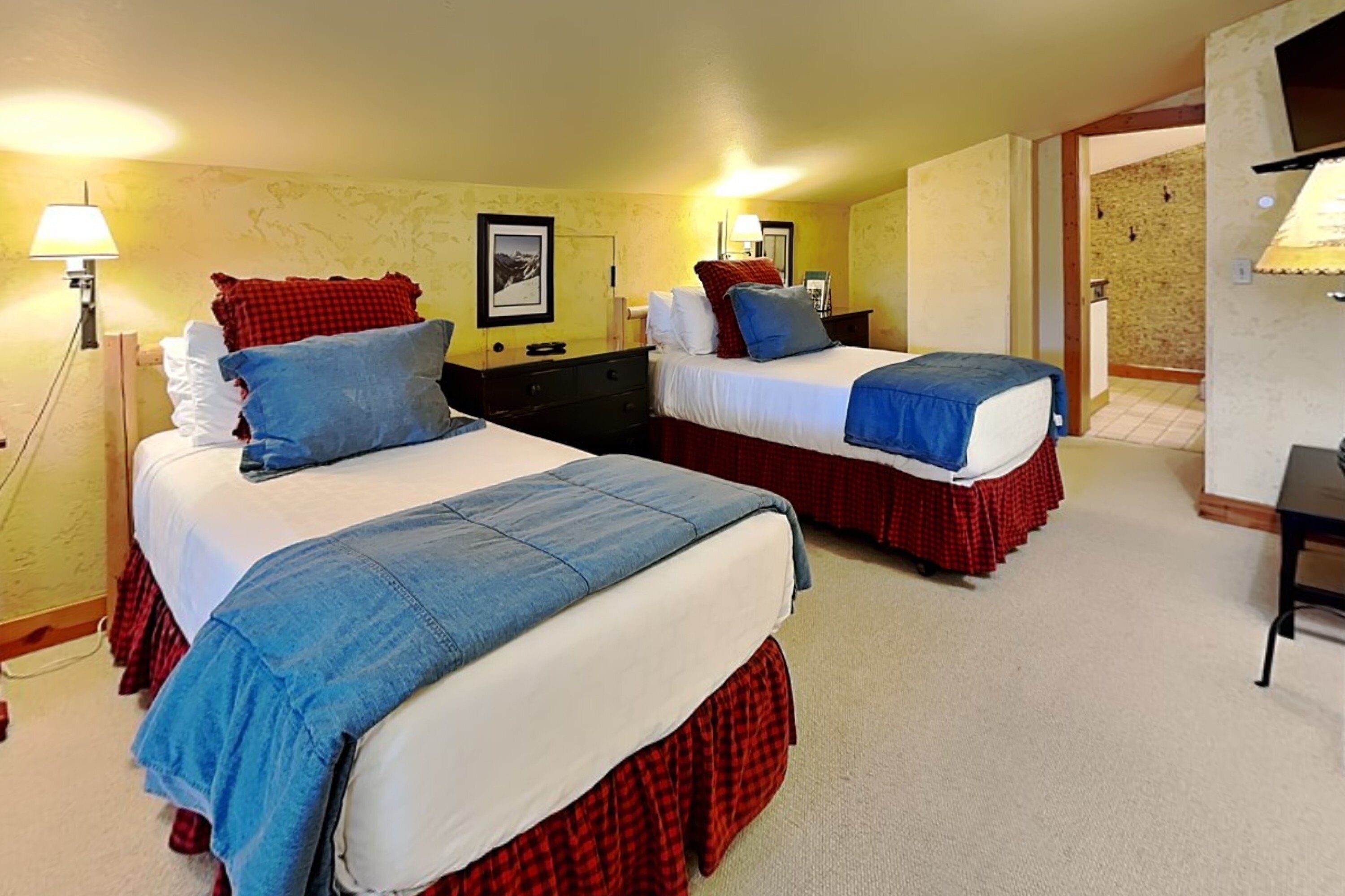 A hotel room with two beds, each with a blue bedspread and red skirted beddings. There are bedside tables with lamps, a wall-mounted TV, and a door leading to another room.