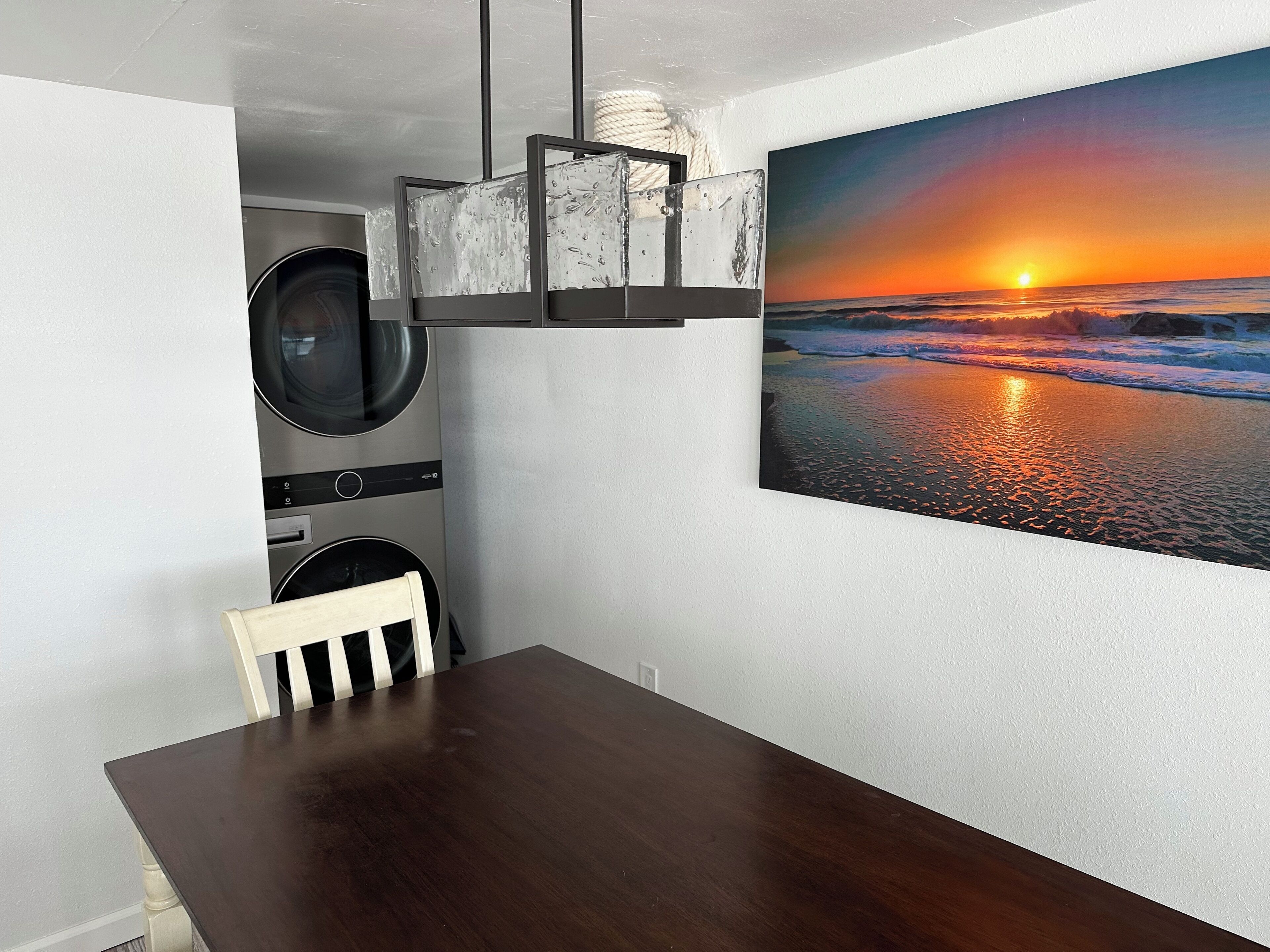 A dining area with a wooden table, a washing machine, and a large painting of a sunset over the ocean.