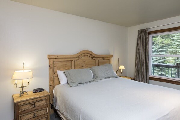 Une chambre à coucher avec une tête de lit en bois, un lit recouvert de linge de lit blanc, deux lampes de chevet et une fenêtre donnant sur des arbres.