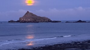 A coastal scene with a prominent rock formation in the center, surrounded by calm sea and a clear sky.