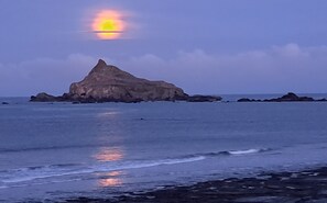 Escena costera con una formación rocosa destacada en el centro, rodeada de mar en calma y un cielo despejado.