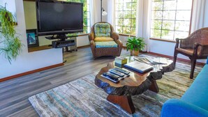 A living room with a television, two wicker chairs, a coffee table with remote controls, and a large window with curtains.