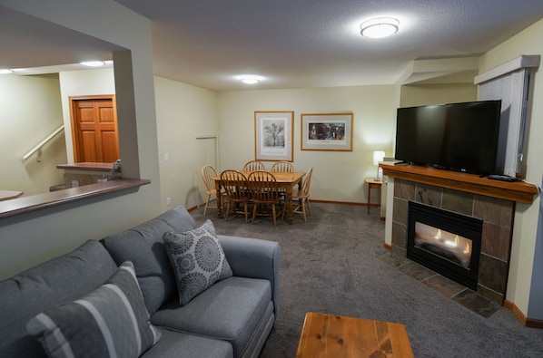 A living room with a gray sofa, a wooden coffee table, a fireplace, a television, a dining table with chairs, and framed pictures on the wall. - Stoney Creek Lagoons 32 (Whistler)