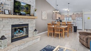 A modern living room with a stone fireplace, dining area, kitchen, and a ceiling fan.