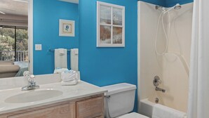 A bathroom with a sink, toilet, and shower. There are towels and a framed picture on the wall.