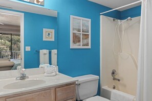 A bathroom with a sink, toilet, and shower. There are towels and a framed picture on the wall.